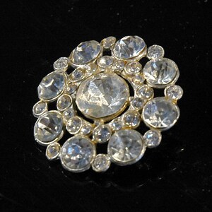 May include: A silver-toned button with a circular design featuring a cluster of clear rhinestones of varying sizes.