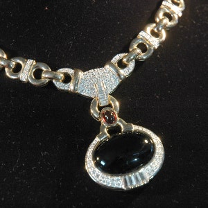 Puede incluir: Un collar de oro con un colgante ovalado negro engastado con diamantes y una piedra roja. El collar tiene un diseño de cadena de eslabones con detalles de diamantes.