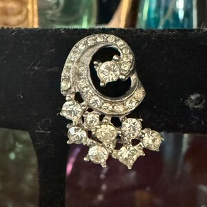 May include: A silver-toned earring with a swirling design, encrusted with clear, sparkling rhinestones. The earring features a cluster of larger stones at the bottom, adding a touch of glamour. The overall design is elegant and vintage-inspired.