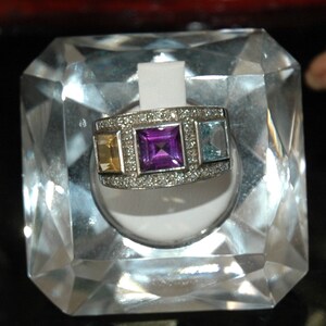 Peut inclure: Une bague en argent avec un sertissage de pierres précieuses violettes, bleues et jaunes. La bague est entourée de petits diamants et est exposée sur un support en cristal transparent.