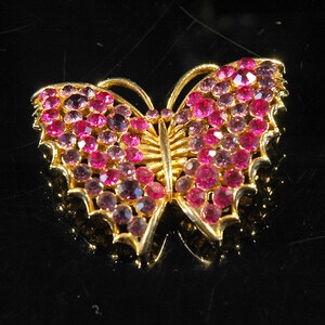 Op de afbeelding: Een gouden vlinder broche met roze strass steentjes. De vlinder heeft een gedetailleerd ontwerp met ingewikkelde vleugels en een lichaam.
