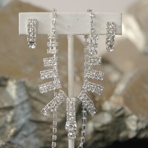 May include: A silver rhinestone necklace and earrings set. The necklace features a long, dangling design with multiple rows of rhinestones. The earrings are simple and feature a single row of rhinestones.