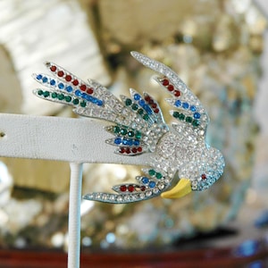 May include: A silver bird brooch with clear, red, blue, and green rhinestones. The bird has a gold beak and is perched on a white stand.