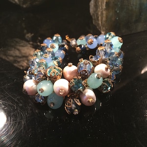 Puede incluir: Un delicado brazalete hecho de cuentas de vidrio azul y blanco, perlas y detalles dorados. El brazalete está diseñado en un estilo de racimo, con las cuentas dispuestas en un patrón suelto y orgánico.