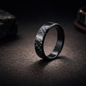 Könnte beinhalten: Ein kreisförmiger Ring mit einem dunkelgrauen und schwarzen Karomuster aus Kohlefaser. Das Band hat eine glatte, polierte Oberfläche mit einem flachen Profil und einer gleichmäßigen Breite über den gesamten Umfang des Schmuckstücks.