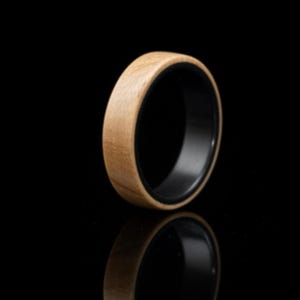May include: A ring featuring a light brown, natural wood grain exterior band paired with a smooth, solid black interior lining. The band has a rounded profile and a polished finish, showcasing the contrast between the organic wood and dark material.