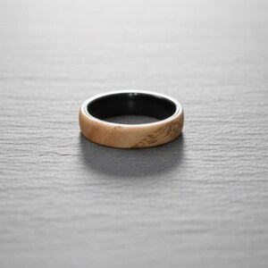 以下が含まれることがあります： 明るいタンカラーの木目調の外側バンドと、滑らかで無地の黒い内側ライニングを備えたリング。バンドは丸みを帯びたプロファイルと磨き上げられた仕上げで、表面全体に木目の自然な質感と模様がはっきりと現れています。