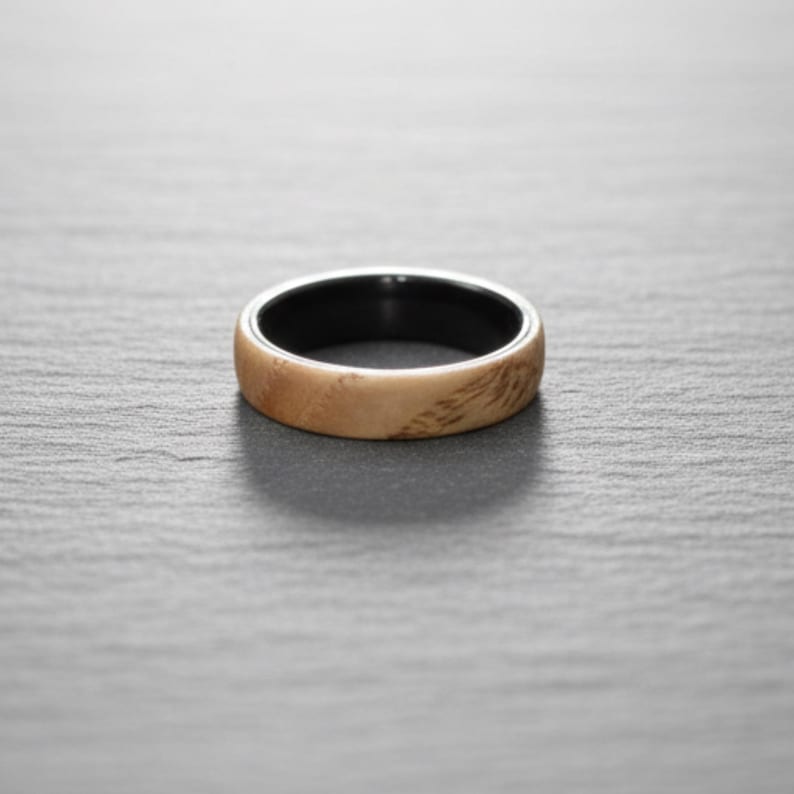以下が含まれることがあります： 明るいタンカラーの木目調の外側バンドと、滑らかで無地の黒い内側ライニングを備えたリング。バンドは丸みを帯びたプロファイルと磨き上げられた仕上げで、表面全体に木目の自然な質感と模様がはっきりと現れています。