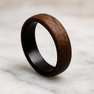 Könnte beinhalten: Ein Holzring mit dunkelbrauner Außenseite und schwarzer Innenseite. Der Ring hat eine glatte, polierte Oberfläche, die die natürliche Holzmaserung zur Geltung bringt. Der Ring ist ein schlichtes, elegantes Design.