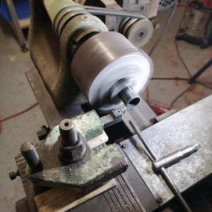 以下が含まれることがあります： 緑色の鋳鉄製フレームを備えた金属旋盤で、回転するチャックが円筒形の金属ワークピースを保持しています。切削工具がツールポストに取り付けられており、T字型のチャックハンドルが機械の平らなベッドの上に置かれています。