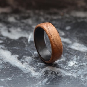 K&ouml;nnte beinhalten: Ein Holzring mit dunklem Innenraum. Der Ring besteht aus hellbraunem Holz mit sichtbarer Maserung. Der Ring liegt auf einer grau-wei&szlig;en Marmoroberfl&auml;che.