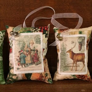 Puede incluir: Cuatro adornos navideños con imágenes vintage de Papá Noel y escenas invernales. Cada adorno de tela tiene un lazo de cinta para colgar y está decorado con pequeñas campanas o bayas rojas. Los adornos están sobre una superficie de madera.
