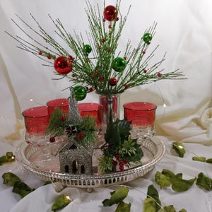 May include: A silver tray with a red and green Christmas centerpiece. The centerpiece includes a silver church, red and green ornaments, and a sprig of holly. The tray is surrounded by green leaves.
