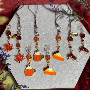 May include: A collection of autumn-themed charms. The charms feature maple leaves, pumpkin spice lattes, and pumpkin pie slices. Each charm is adorned with orange and red beads and a black cord.
