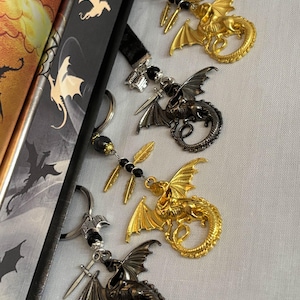 Puede incluir: Cuatro marcapáginas de dragón con lazos de cinta negra. Cada marcapáginas presenta un dragón con una espada y plumas. Los dragones son dorados, negros y plateados.