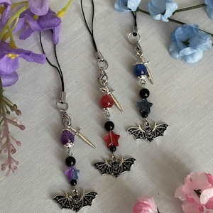 Może przedstawiać: Trzy breloczki do telefonu z czarnymi nietoperzami, srebrnymi mieczami i kolorowymi koralikami z kamieni szlachetnych. Każdy breloczek ma czarny koralik w kształcie gwiazdy i srebrną pętelkę do przymocowania do etui na telefon.