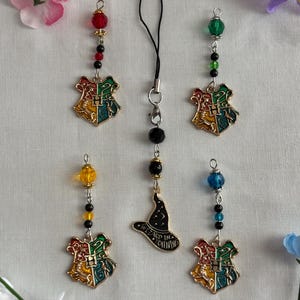 Könnte beinhalten: Fünf dekorative Anhänger mit dem Hogwarts-Wappen und einem Zaubererhut. Jeder Anhänger hat ein goldfarbenes Wappen mit roter, grüner, gelber und blauer Emaille. Die Anhänger sind mit bunten Perlen und einem schwarzen Schlüsselband verziert.