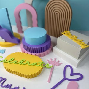 May include: A collection of colorful, decorative cake toppers and props. Includes a yellow "Celebrate" sign, a pink crown, and purple and pink heart-shaped wands. Also features a multi-tiered cake with blue, purple, and pink layers.