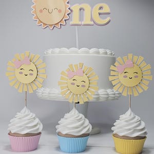 Può includere: Una torta bianca glassata con un cake topper "one" e tre decorazioni per cupcake. Le decorazioni presentano disegni di sole sorridenti con fiocchi rosa. I cupcake hanno glassa bianca e sono in pirottini rosa, blu e gialli.