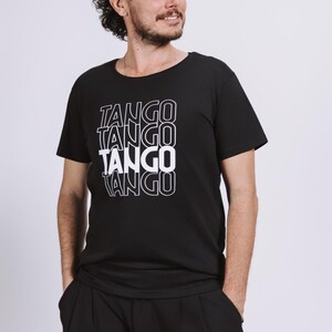 以下が含まれることがあります： 黒の半袖Tシャツで、TANGOという単語が縦に4段重ねてプリントされています。テキストは白いアウトラインフォントで描かれており、衣服の胸の中央に繰り返されるグラフィックデザインが施されています。