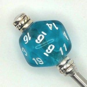May include: A teal blue, glass D20 die bead with white numbers, attached to a silver-colored metal stick. The die has the numbers 3, 6, 8, 9, 11, 14, 16, and 19 visible. The top and bottom of the die are capped with decorative silver metal.
