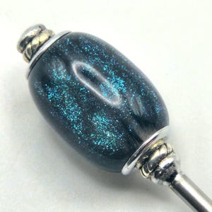 May include: A decorative bottle stopper with a dark blue, glittery, oval-shaped body. The stopper has silver-colored metal accents at the top and bottom, with a twisted rope design on the bottom. The top has a small gold-colored accent.