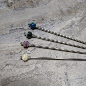 May include: Four silver stirring sticks with different colored glass beads on the end. The beads are green, white, blue, and multi-colored.