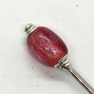 May include: A decorative bottle stopper with a red, oval-shaped bead. The bead has a glittery, speckled appearance and is capped with silver-colored metal. The stopper has a long, slender, silver-colored handle.