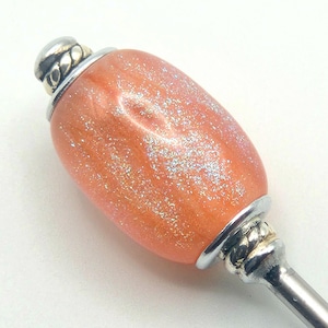 May include: A decorative bottle stopper with a pearlescent orange body and silver accents. The stopper has a glittery finish and a silver metal base and top. The top features a decorative silver rope design.