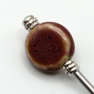 Puede incluir: Un tapón de botella decorativo con una parte superior ovalada de cerámica granate y crema. La parte superior tiene una apariencia texturizada y está acentuada con detalles de metal plateado. El tapón tiene un vástago de metal plateado.