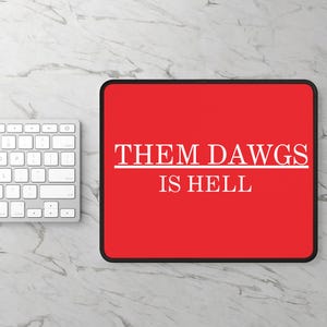 Puede incluir: Alfombrilla de ratón rectangular roja con el texto blanco "THEM DAWGS IS HELL". La alfombrilla tiene un borde negro y está junto a un teclado blanco sobre una superficie con vetas de mármol.
