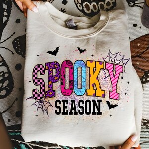 May include: A cream-colored sweatshirt with the words "SPOOKY SEASON" in colorful, patterned letters. The design includes bats, spiderwebs, and stars, creating a festive Halloween theme. The sweatshirt is folded.