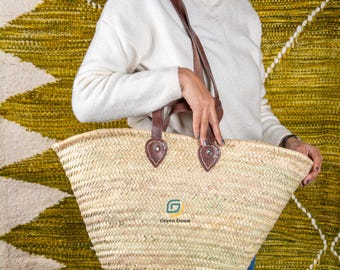 May include: A woven straw tote bag with brown leather handles and accents. The bag features a wide, open design and a green logo that reads "Green Doum". The bag is held by a person wearing a white sweater.