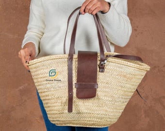 May include: Woven straw tote bag with brown leather handles and a matching flap closure. The bag features the text "Green Doum" and a green logo. The bag is held by a person.