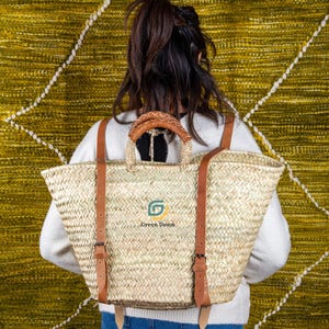Puede incluir: Mochila de paja tejida con correas y asa de cuero marrón. La bolsa tiene forma rectangular con una abertura ancha. El texto "Green Doum" y un logotipo verde son visibles en la parte delantera de la bolsa.