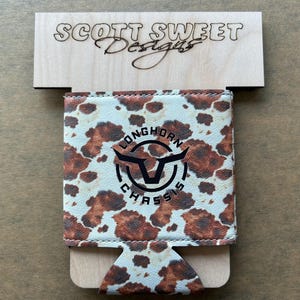 May include: A brown and white cowhide-patterned can cooler with the text "LONGHORN CHASSIS" and a longhorn logo. The cooler is displayed on a wooden stand with the text "SCOTT SWEET Designs" etched into it.