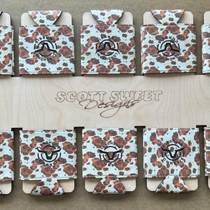 May include: A display of ten can coolers with a cowhide pattern. Each cooler features a black logo with a longhorn and the words "Longhorn Classic". The wooden display board reads "Scott Sweet Designs".