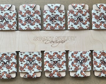 May include: A display of ten can coolers with a cowhide pattern. Each cooler features a black logo with a longhorn and the words "Longhorn Classic". The wooden display board reads "Scott Sweet Designs".