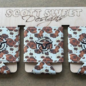 May include: Three Longhorn Chassis can coolers with a cowhide pattern, displayed on a wooden holder. Each cooler features the Longhorn Chassis logo in black and white. The wooden holder has the text "SCOTT SWEET Designs" engraved.