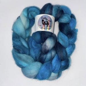 Könnte beinhalten: Ein Strang blaue Kunstfaser, mit Schattierungen von hell- und dunkelblau. Ein weißes Etikett mit dem Text "Curly Ewes Fiber Arts" und einem Schaf-Logo ist um das Garn gewickelt.
