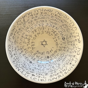 May include: A white ceramic bowl featuring black hand-written Hebrew text spiraling from the rim towards the centre. The interior base displays a small, centred Star of David symbol, with additional smaller Star of David icons interspersed throughout.