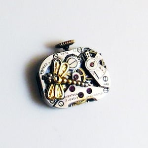 May include: A silver watch movement with a gold dragonfly design. The watch face is labeled "Seventeen Jewels" and "Swiss Made".