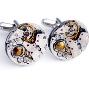 May include: A pair of silver cufflinks with a circular design featuring exposed watch gears and small purple gemstones.