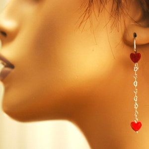 May include: Silver dangle earrings with red heart charms. The earrings are hanging from the earlobe of a mannequin.