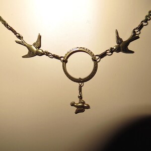 May include: A gold necklace with three bird charms. The necklace has a circular pendant with a decorative design. The birds are in a flying position.