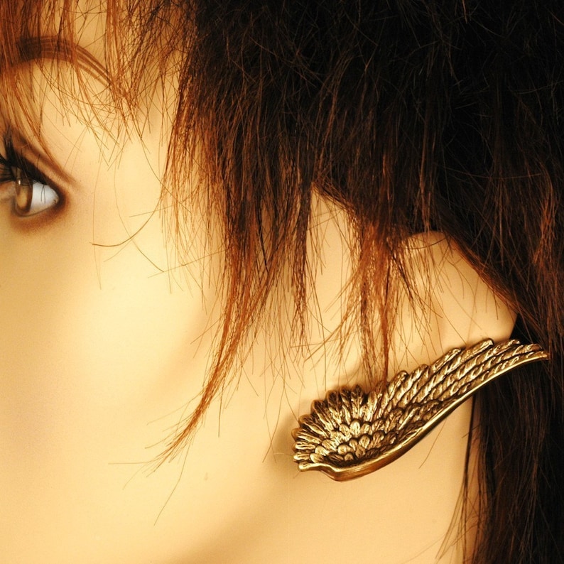 Pode incluir: Um brinco dourado em forma de asa com um design detalhado. O brinco &eacute; usado na orelha esquerda de uma pessoa com cabelo castanho.