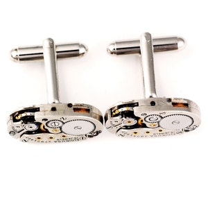Pode incluir: Um par de bot&otilde;es de punho de prata com um design de movimento de rel&oacute;gio em tom dourado. Os bot&otilde;es de punho apresentam o texto "ELGIN USA 15 JEWELS" na face.