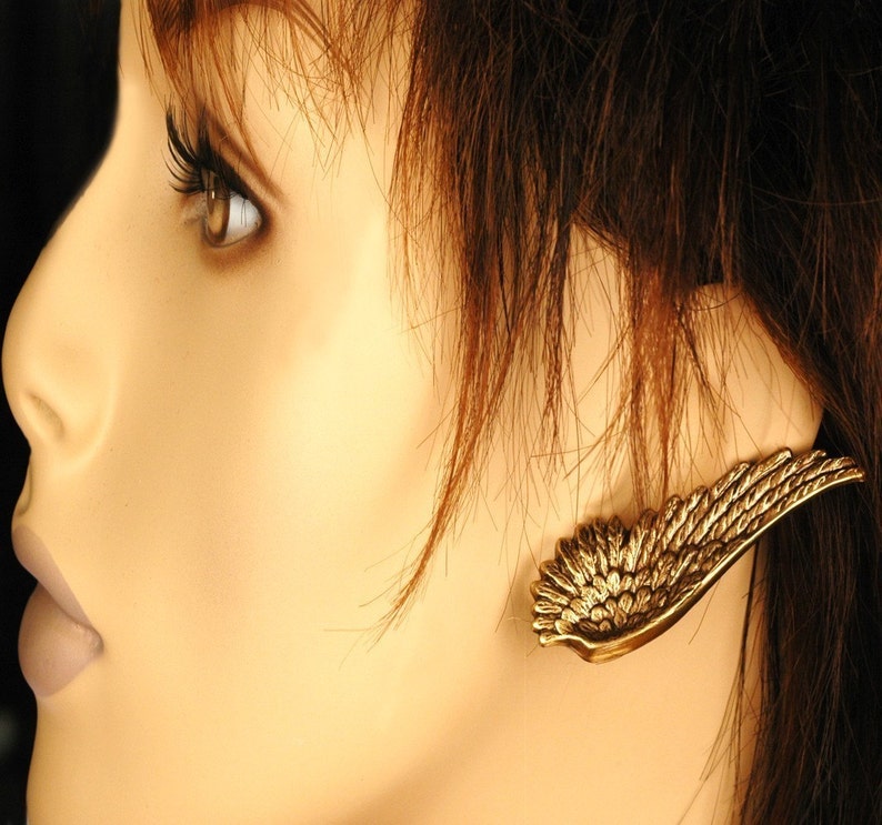 Pode incluir: Um brinco dourado em forma de asa com um design detalhado. O brinco &eacute; usado na orelha de um manequim com cabelo castanho.
