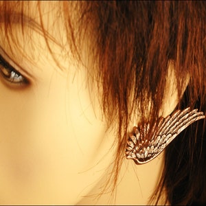 May include: A close-up of a woman's ear with a gold-toned wing earring. The earring is a single wing with a detailed design.