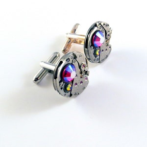 May include: A pair of silver cufflinks with a circular design. Each cufflink features a small, iridescent red jewel in the center, surrounded by a silver gear mechanism.